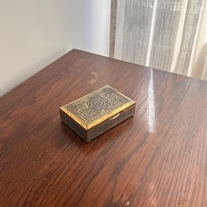 Op de afbeelding: Een rechthoekige metalen doos met een ingewikkeld donkerblauw en goudkleurig filigraanpatroon op het deksel en de zijkanten. De doos heeft een scharnierend deksel met een kleine gouden sluiting aan de voorzijde en gedetailleerde gravures.