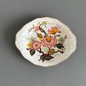Op de afbeelding: Een ovale keramische schaal met een geschulpte rand en gouden afwerking. Het midden toont een kleurrijk bloemmotief met roze, gele en blauwe bloemen, groene bladeren en donkere stelen op een crèmekleurige achtergrond met een subtiele craquelé.