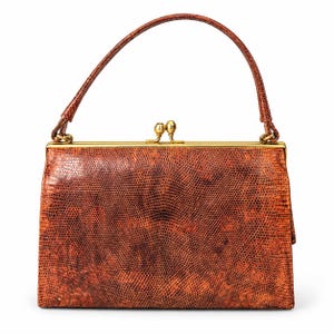 Könnte beinhalten: Eine strukturierte Handtasche mit einer texturierten, braun-orangen Eidechsenhaut-Optik. Die Tasche verfügt über einen einzelnen Tragegriff, einen goldfarbenen Metallrahmen und einen Verschluss mit zwei Knöpfen an der Oberseite. Die Oberfläche zeigt ein feines Muster.