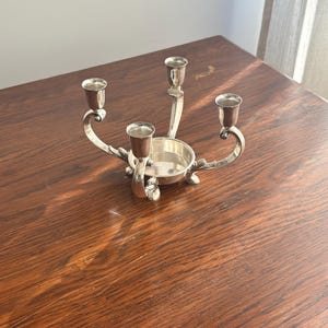 May include: A silver-colored metal candelabra featuring three candle holders arranged around a central circular base. The structure has curved arms with decorative accents at the base of each holder, providing a polished, reflective surface.