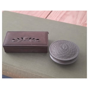 Op de afbeelding: Twee antieke metalen dozen. Eén is rechthoekig met een bloemenontwerp en een scharnierend deksel. De andere is rond met een gestructureerd, patroonvormig deksel. Beide zijn in tinten bruin en grijs.