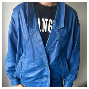 Op de afbeelding: Een felblauwe leren jas met een klassieke reverskraag en een sluiting met één knoop. De jas heeft een getailleerde pasvorm met zakken en een gestikt ontwerp. De persoon draagt een zwart t-shirt met het woord "ANG" in het wit.