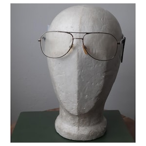 Puede incluir: Gafas de sol estilo aviador con una fina montura de metal dorado y lentes transparentes. Las gafas tienen un doble puente y un diseño clásico. Las almohadillas nasales son de color marrón claro. Las gafas se exhiben en una cabeza de espuma blanca.