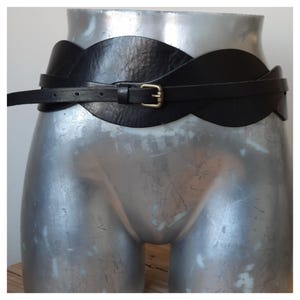 Op de afbeelding: Een zwarte leren riem met een breed, golvend ontwerp en een dunne riem. De riem heeft een goudkleurige gesp en wordt op een mannequin getoond. Dit is een modeaccessoire.