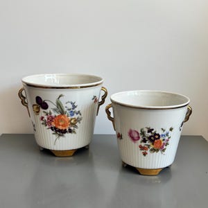 Op de afbeelding: Twee witte porseleinen plantenpotten met verticale ribbels, voorzien van kleurrijke bloemenboeketten aan de voorzijde. Elke pot heeft goudkleurige handvatten en goudkleurige voetjes. De grotere pot staat links en de kleinere staat rechts.