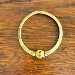 Op de afbeelding: Een goudkleurige omega-ketting met een centrale ronde hanger. De hanger is versierd met drie kleine zwarte stenen, één kleine rode steen en twee heldere accentstenen die aan weerszijden van het centrale ronde element zijn geplaatst.