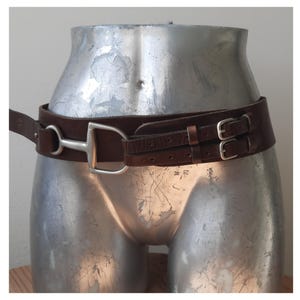 Op de afbeelding: Een bruine leren riem met een zilveren bit en gespsluiting. De riem is om een zilveren mannequin torso gewikkeld. De riem heeft een breed ontwerp en is waarschijnlijk voor gebruik in de paardensport.