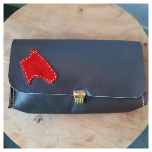 Op de afbeelding: Een donkergrijze leren clutch met een rode paardenkop-applicatie. De clutch heeft een goudkleurige sluiting en is gestikt met oranje draad. De rechthoekige tas lijkt handgemaakt.