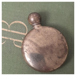 Op de afbeelding: Een ronde, metalen zakfles met een verweerde, bronskleurige afwerking en een bolvormige schroefdop. Het oppervlak van de fles vertoont zichtbare krassen en gebruikssporen, met een glad, rond lichaam en een korte, smalle hals.