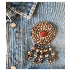 Op de afbeelding: Zilverkleurige broche met een centrale rode steen en bungelende kralen. De ronde broche is geplaatst op een spijkerjasje, wat het gedetailleerde ontwerp en de vintage stijl benadrukt. Het ingewikkelde metaalwerk van de broche voegt elegantie toe.