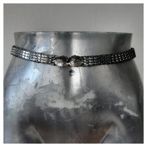 Op de afbeelding: Een zilverkleurige riem met een blad-vormige sluiting. De riem is gemaakt van kleine, met elkaar verbonden rechthoekige stukjes. De sluiting is een paar gestileerde bladeren. De riem wordt getoond op een zilveren mannequin.