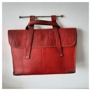 Puede incluir: Un maletín de cuero rojo con asa y bandolera, colgado de una barra de metal. El maletín tiene una superficie texturizada y costuras visibles. La bolsa es de estilo vintage.