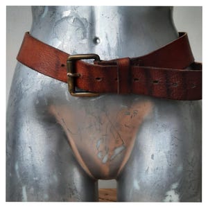 Pode incluir: Um cinto de couro castanho com uma fivela retangular de latão. O cinto está enrolado em volta de um torso de manequim prateado. O couro mostra sinais de desgaste, aumentando o seu apelo vintage. Um acessório clássico.