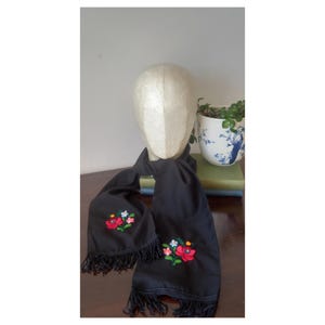 Op de afbeelding: Zwarte sjaal met geborduurde bloemmotieven in rood, roze en blauw. De sjaal heeft een gefranjerde rand en is over een mannequinhoofd gedrapeerd. Een decoratieve bloempot staat op de achtergrond.
