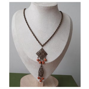 Può includere: Una collana vintage color bronzo con un pendente e catena in filigrana. Il pendente presenta un design a forma di diamante con dettagli intricati e piccole perle arancioni. La collana è esposta su un manichino bianco.