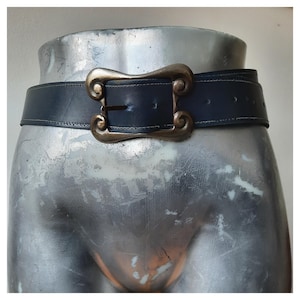 Könnte beinhalten: Ein dunkelblauer Ledergürtel mit einer dekorativen, bronzefarbenen Schnalle. Die Schnalle hat ein verziertes, wirbelndes Design. Der Gürtel wird auf einem Mannequin-Torso präsentiert.