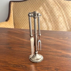 Op de afbeelding: Een zilverkleurige metalen kruidenstandaard met een centrale verticale paal, een ronde voet en een bovenste ring. De standaard bevat een klein mes en een vork met twee tanden die verticaal aan de bovenste ring hangen.