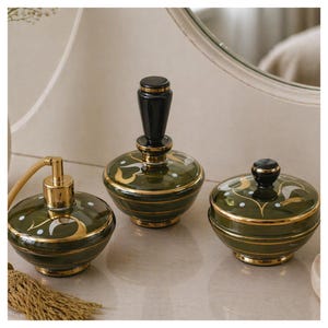 Pode incluir: Um conjunto de três artigos de toucador em cerâmica de estilo vintage. As peças são verde-oliva com detalhes dourados e brancos. O conjunto inclui um frasco de perfume, um pote com tampa e um atomizador.