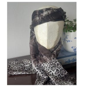 Puede incluir: Una cabeza de maniquí está cubierta con una bufanda con estampado de animales marrón oscuro y blanco y una diadema a juego. La bufanda está envuelta alrededor del cuello y cae. Una pequeña maceta blanca con un diseño floral azul y una planta verde está en el fondo.