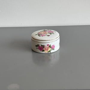 Op de afbeelding: Een klein, rond wit porseleinen doosje met een delicaat bloemmotief van roze, gele en rode rozen met groene bladeren. Het deksel en de basis zijn afgewerkt met een dunne, metallic gouden rand langs de zijkanten voor een verfijnde uitstraling.