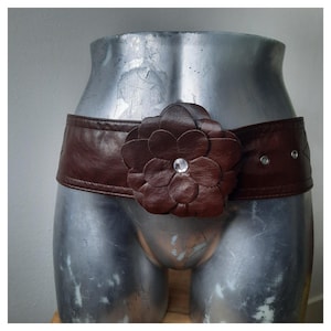 Op de afbeelding: Een bruine leren riem met een grote, bloemvormige versiering. De bloem heeft meerdere bloemblaadjes en een heldere edelsteen in het midden. De riem wordt op een mannequin gepresenteerd.