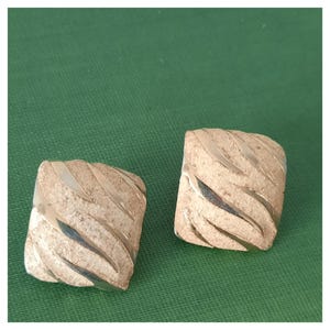 Op de afbeelding: Een paar goudkleurige vierkante oorbellen met een getextureerd, geribbeld ontwerp. De oorbellen hebben een geborstelde metalen afwerking en een afgeronde rand. De oorbellen worden gepresenteerd op een groene achtergrond.