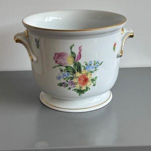 Op de afbeelding: Een witte porseleinen plantenpot met een handgeschilderd bloemenboeket in roze, gele en blauwe tinten. De pot heeft twee zijhandvatten met gouden accenten, een goudkleurige rand aan de bovenkant en een ronde voet met verfijnde details.