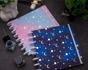 May include: Two disc-bound notebooks with celestial designs. One has a pink and blue gradient with stars and suns, the other a dark blue background with gold moons and stars. White binding and elastic closure.