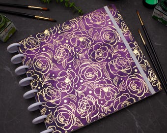 May include: A purple planner with a gold floral design and a white disc binding system. The planner has a white elastic closure.