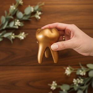 Könnte beinhalten: Ein goldfarbenes, zahnförmiges Dekorationsobjekt. Das Objekt wird in einer Hand vor einem dunklen Holzgrund gehalten, mit Zweigen aus Grün und weißen Blumen im Hintergrund.