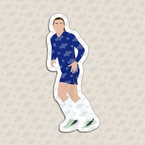 May include: A die-cut sticker featuring a stylized illustration of a soccer player wearing a blue long-sleeve jersey with a geometric pattern, blue shorts, white socks, and light green cleats. The player is depicted in a running pose.