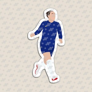 May include: A die-cut sticker featuring a soccer player in a dark blue long-sleeve jersey with a light blue geometric pattern and a yellow logo. The player wears white socks and white soccer cleats with red accents, depicted in a running pose.