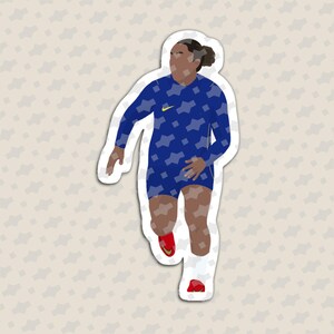 May include: A die-cut sticker featuring a stylized illustration of a soccer player in a blue long-sleeve jersey with a geometric pattern, dark blue shorts, white socks, and red cleats. The player has dark hair tied back and a yellow logo on the chest.