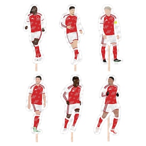 May include: A set of six die-cut paper toppers featuring stylized illustrations of soccer players in red and white uniforms. Each figure is attached to a thin wooden stick, depicting athletes in various poses, including running and standing positions.