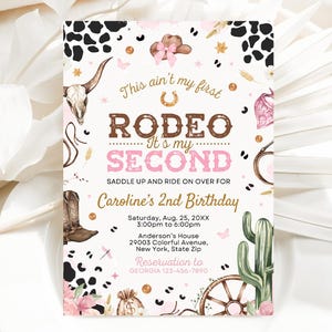 May include: A pink and brown invitation for a second birthday party with a rodeo theme. The invitation features a cow skull, a horseshoe, a cactus, and a wagon wheel. The text on the invitation reads "This ain't my first Rodeo - It's my Second" and "Caroline's 2nd Birthday".