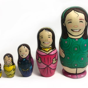 Pode incluir: Um conjunto de cinco bonecas russas de madeira, cada uma pintada com um rosto sorridente e roupas diferentes. As bonecas variam em tamanho, com a maior vestindo um vestido verde e a menor um vestido amarelo. Dispostas em fila sobre um fundo branco.