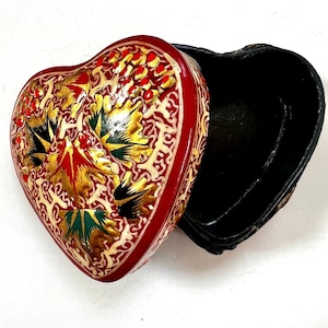 Pode incluir: Uma caixa vermelha em forma de coração com intrincados desenhos florais em ouro e preto. A caixa está aberta e o interior é preto.