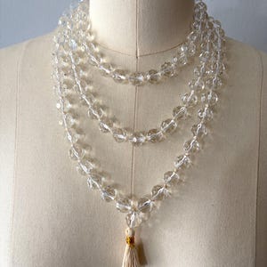 May include: A multi-strand necklace featuring clear, faceted, round beads strung on a light-colored cord. The necklace is arranged in three cascading loops and terminates at the bottom with a small, light-colored fabric tassel with gold thread.