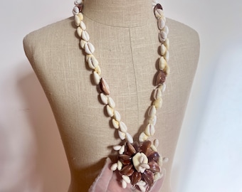 Könnte beinhalten: Eine weiße und braune Muschelkette mit einem großen Muschelblütenanhänger. Die Kette besteht aus Kaurischnecken und ist ein traditionelles hawaiianisches Lei.