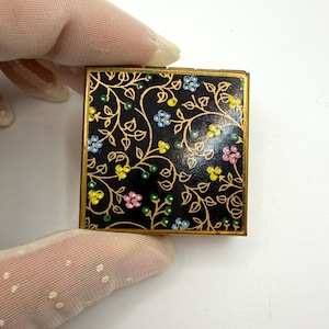 May include: A square compact case with a black background featuring a gold-toned vine pattern and small painted flowers in blue, yellow, pink, and green. The compact has a metallic gold-colored frame and a smooth, flat surface design.
