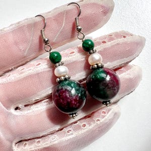 Puede incluir: Un par de pendientes colgantes con grandes cuentas redondas en tonos rojo oscuro y verde. Los pendientes también incluyen cuentas verdes y blancas más pequeñas y ganchos plateados.