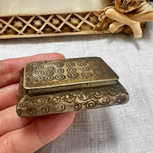 Puede incluir: Una pequeña caja de joyería rectangular de color plateado con un diseño ornamentado. La caja tiene una tapa con bisagras y un patrón decorativo de círculos y líneas.