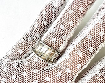 May include: A silver ring with a wide band and a chain design. The ring is worn on a hand covered by a white lace glove. The ring's design features vertical and horizontal chain links, creating a textured appearance.
