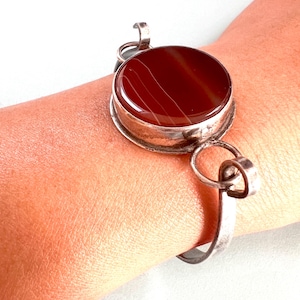 May include: A silver bracelet featuring a large, round, red agate stone. The stone has white lines running through it. The bracelet has a simple, elegant design.
