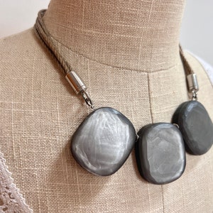 Peut inclure: Un collier ras de cou argenté et gris avec trois gros éléments carrés facetés. Le collier est fait d'un cordon épais en forme de corde.