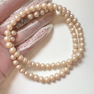 Puede incluir: Un collar de perlas de doble hebra con un cierre plateado. Las perlas son de un color crema suave y parecen de tamaño uniforme. El collar se muestra sobre una superficie rosa y con encaje.