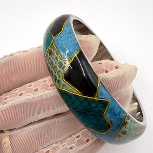 May include: A chunky bangle bracelet with a silver-coloured metal band. The bracelet features a mosaic design in black, blue, and teal, with gold-coloured outlines. A striking fashion accessory.
