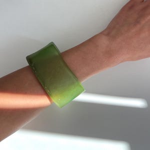 Puede incluir: Un brazalete verde translúcido de forma rectangular. El brazalete se lleva en la muñeca de una persona. Tiene una superficie lisa y un ligero degradado de color, que va de un verde más claro a un tono más oscuro.