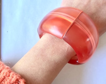 May include: A translucent orange bangle bracelet with a rounded, segmented design. The bracelet has a smooth, glossy finish and is worn on a wrist. The bracelet is a bold statement piece.