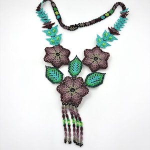 Puede incluir: Un collar de cuentas con tres flores de cinco pétalos de color morado oscuro, detalles en forma de hoja en verde y turquesa, y un fleco de cuentas colgantes en tonos morado, verde y gris. La cadena está formada por pequeñas cuentas moradas.
